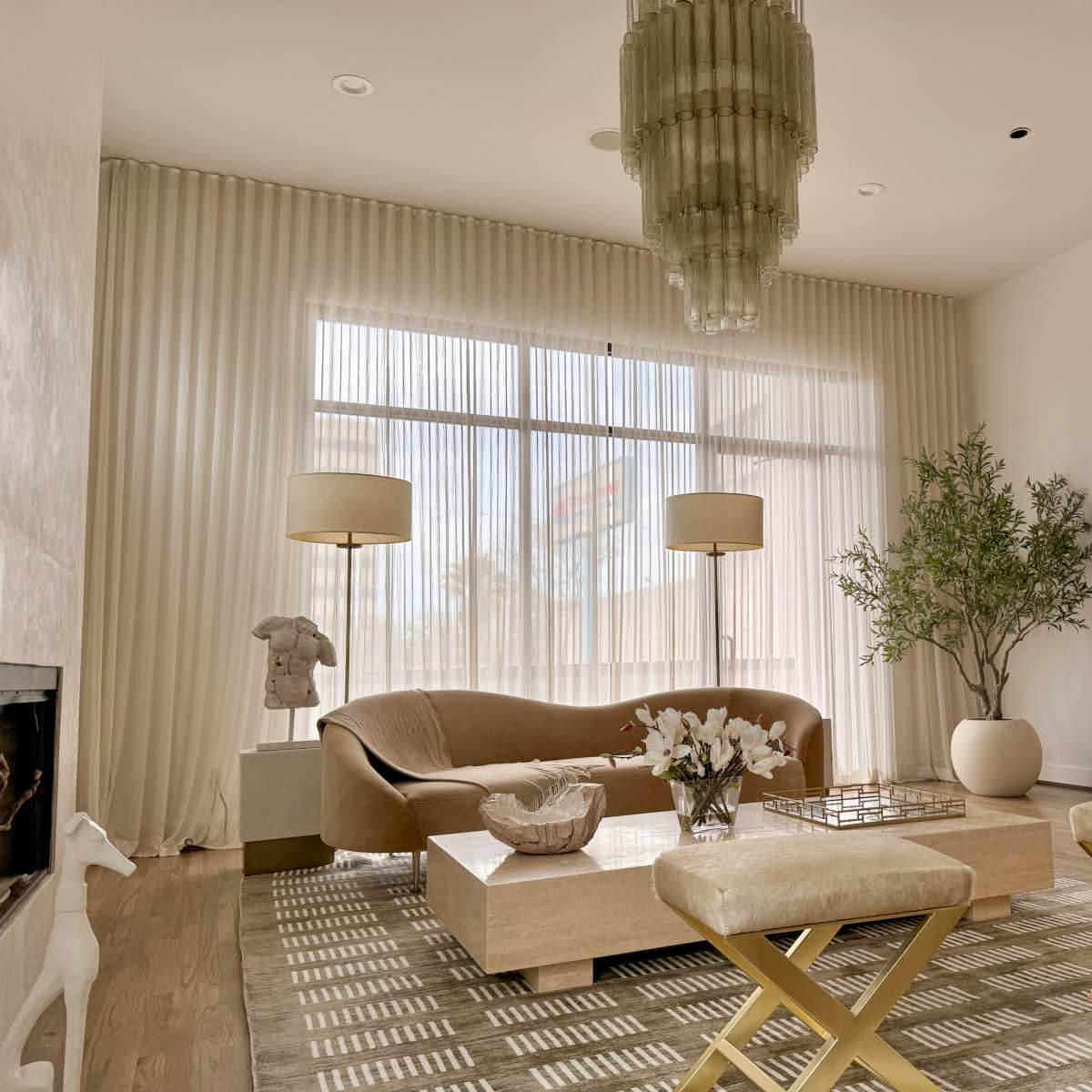 A modern living room features a curved sofa, a coffee table with decorative items, two ottomans, and large windows with sheer curtains that allow natural light.
