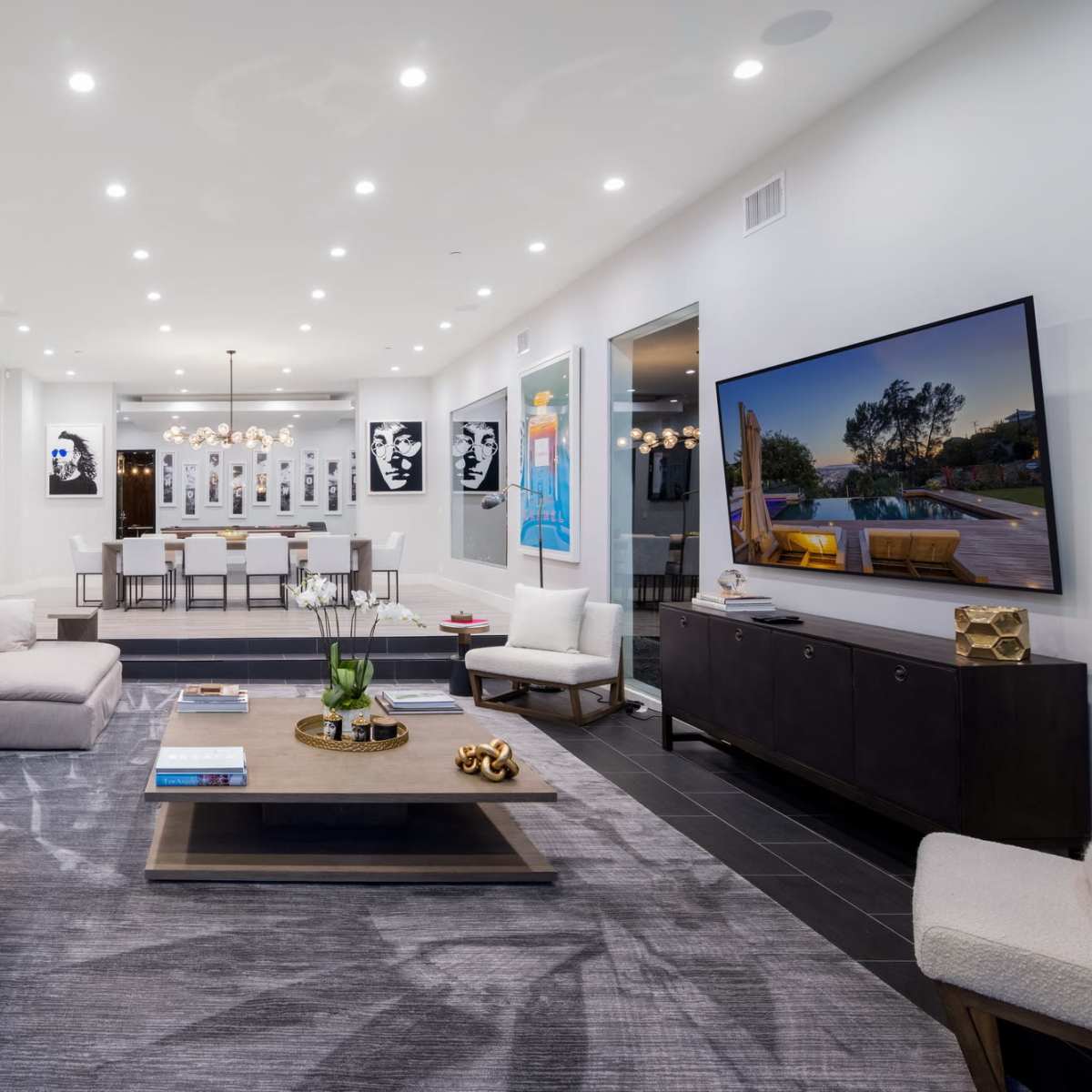A modern living room with a large sectional sofa, a center coffee table, a TV mounted on the wall, and a dining area in the background, decorated with artwork and minimalist decor.