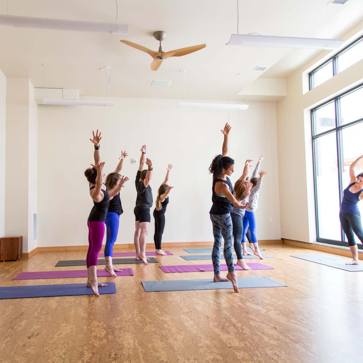 A group of people practices yoga in a spacious studio with large windows and wooden flooring.
