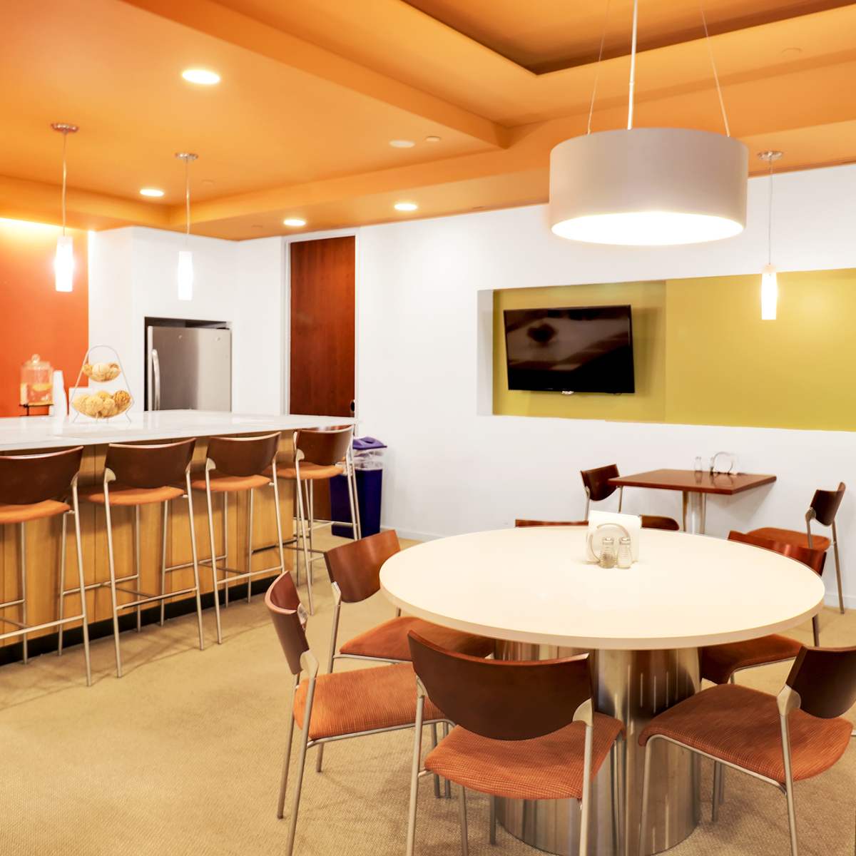 A modern kitchen area with a circular table and wooden chairs, a bar counter with stools, and a wall-mounted television.
