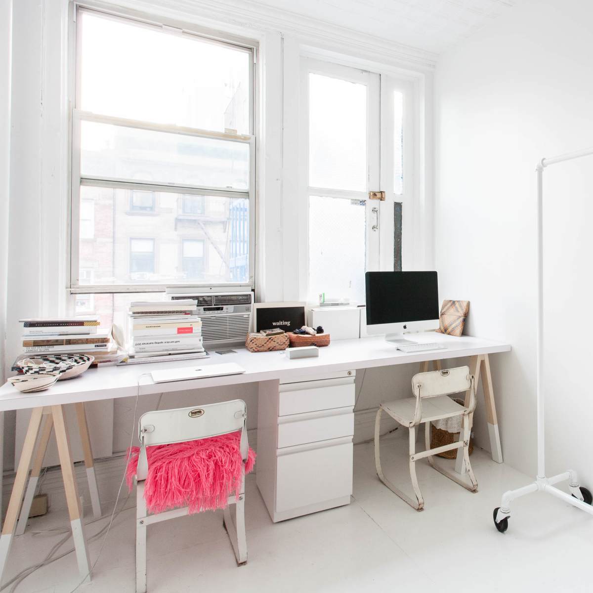 A minimalistic workspace features a white desk with an iMac, stacks of magazines, and two chairs, alongside a clothing rack displaying various garments.