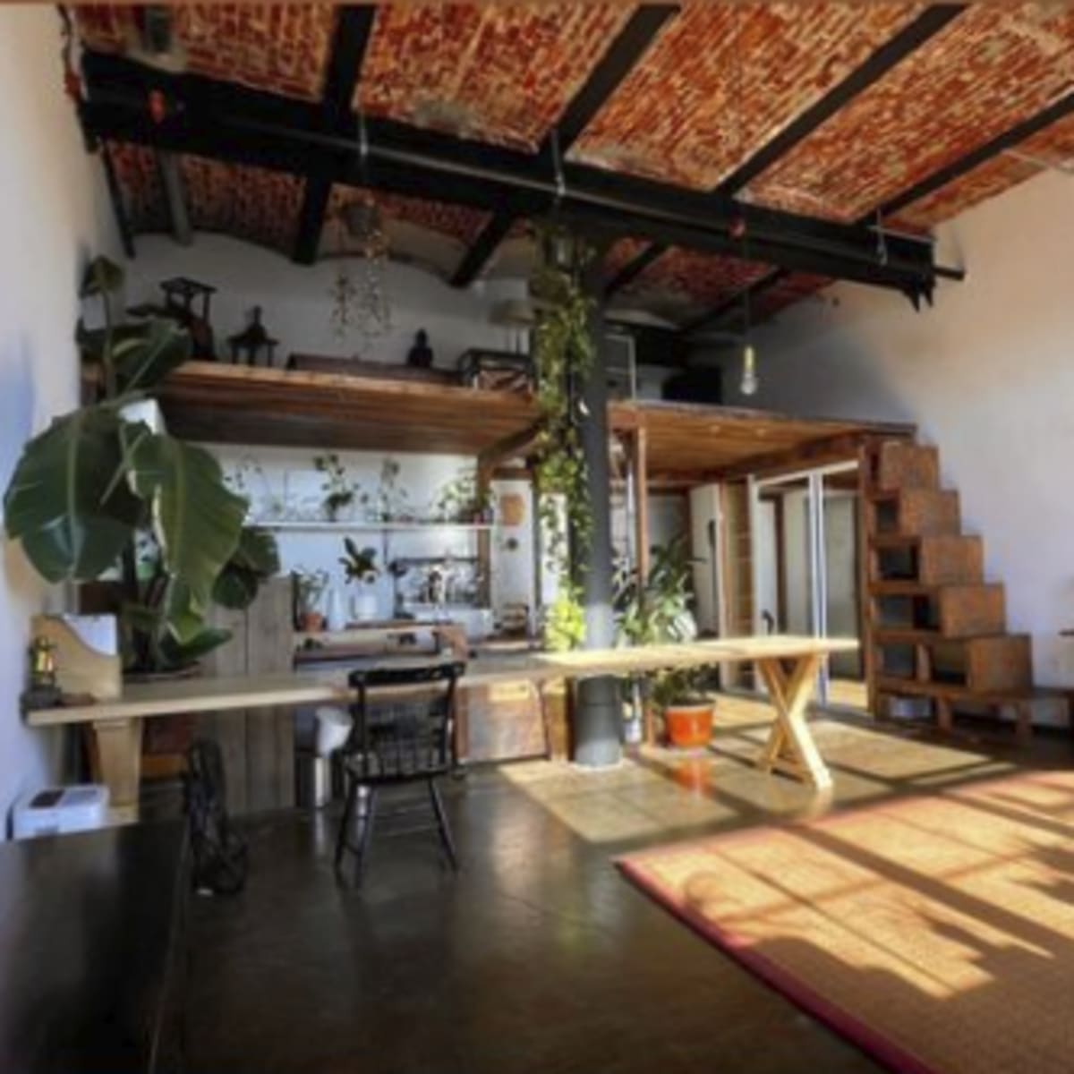 An interior space with exposed brick walls, wooden beams, a staircase, plants, and a living area featuring a large rug and a sofa.