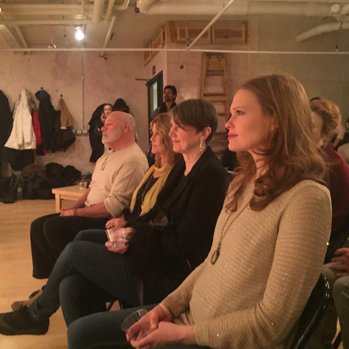 A group of five people sits in chairs, watching a performance in a well-lit room with bare walls and a wooden floor.
