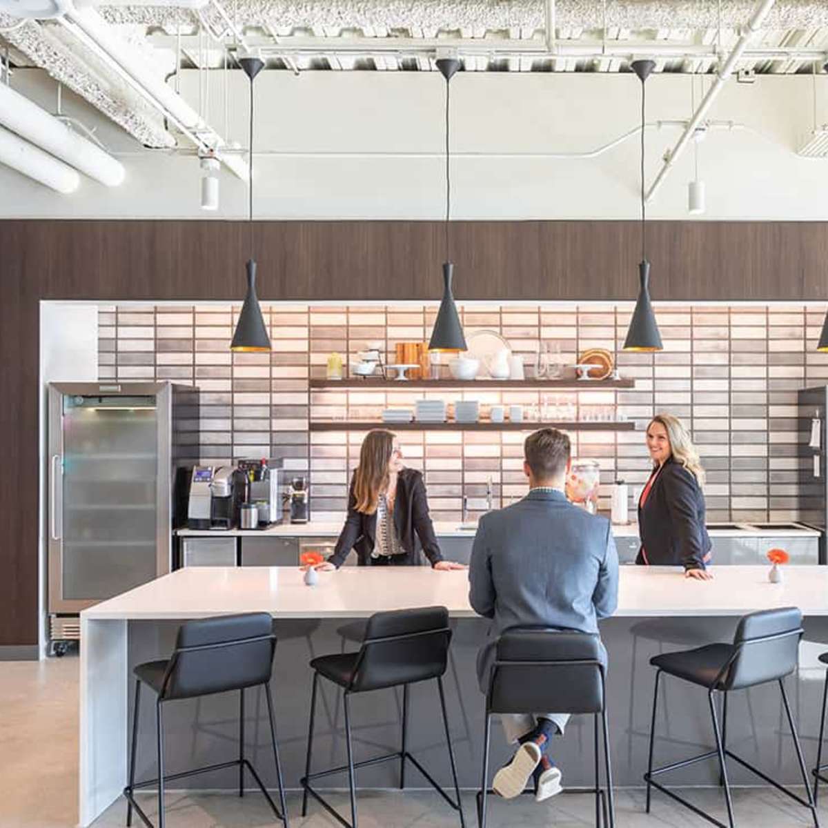 Two women and a man converse at a modern kitchen island in an office setting, surrounded by sleek cabinetry and appliances.