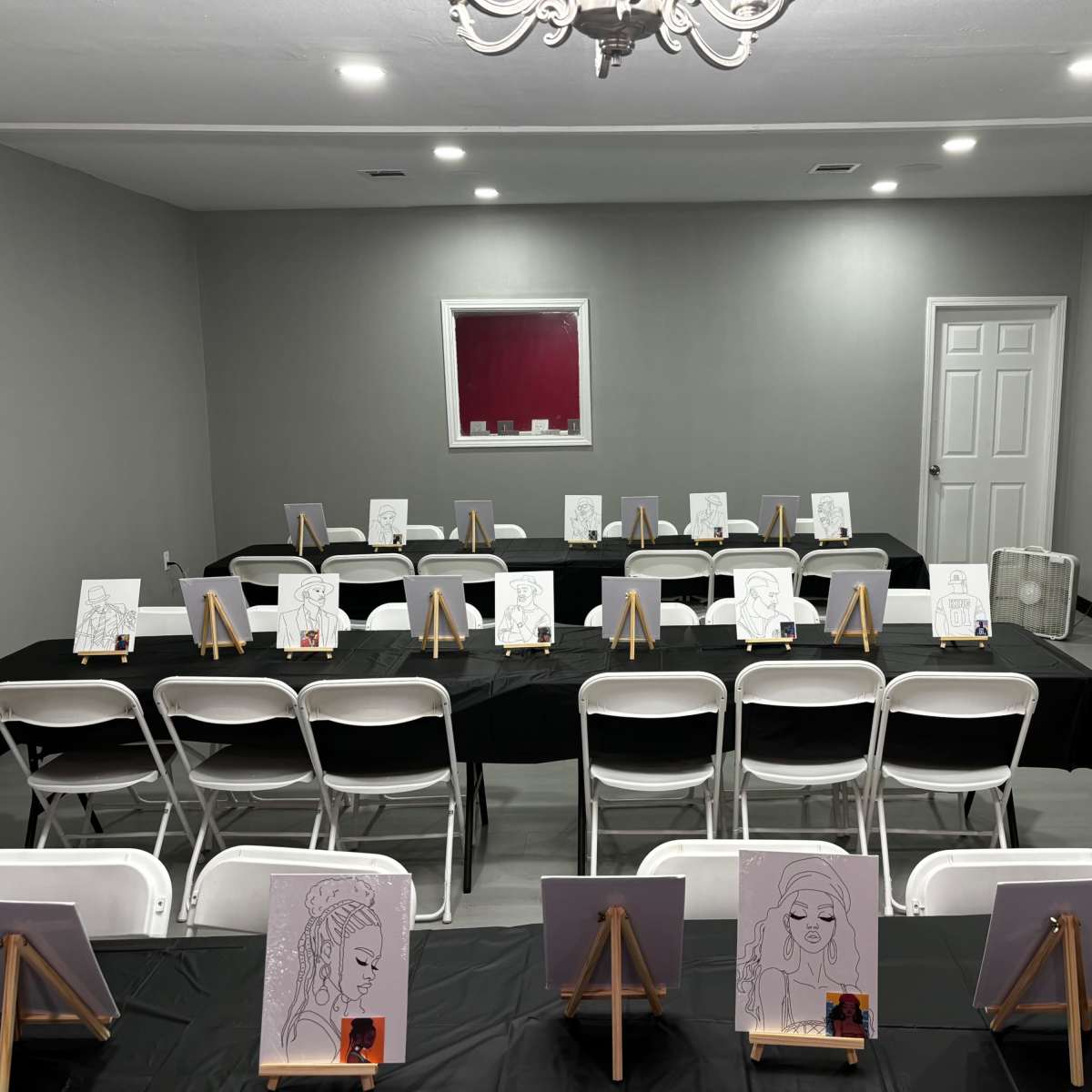 The room features rows of white chairs facing easels with black and white sketches on each, arranged on tables covered with black cloth.