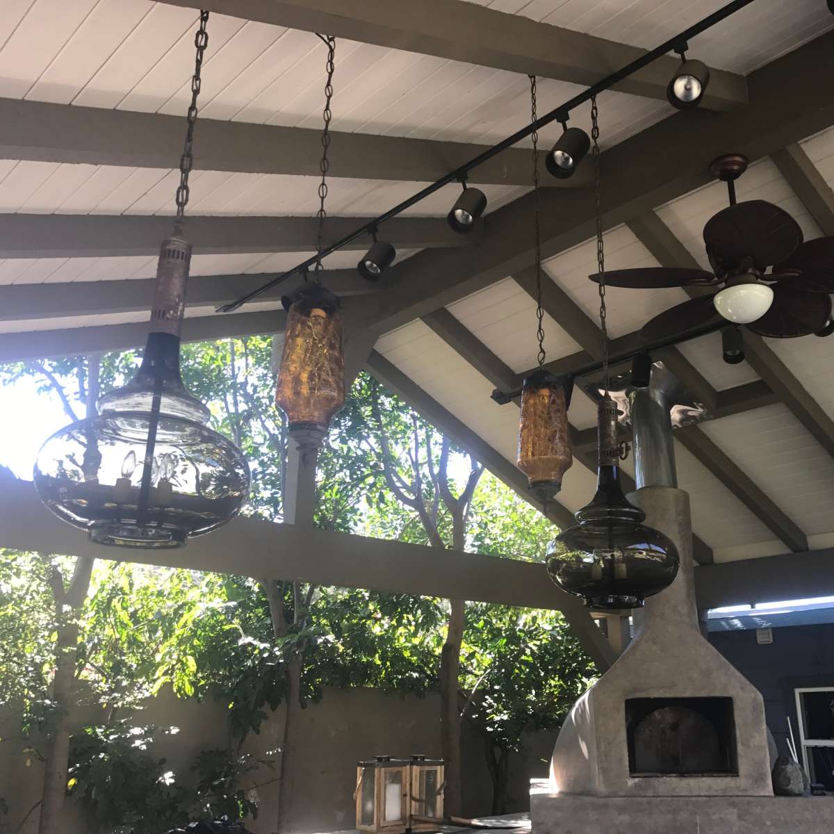 A covered outdoor area with hanging light fixtures and a ceiling fan, surrounded by greenery.