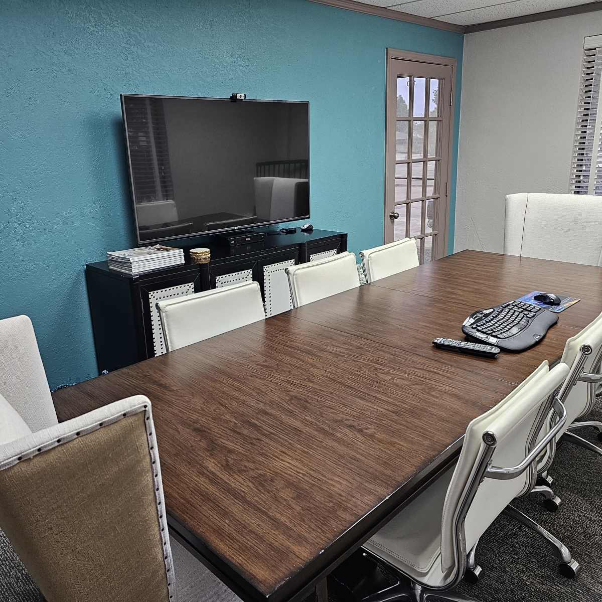 A modern conference room features a long wooden table surrounded by white ergonomic chairs, with a large television mounted on a blue wall.