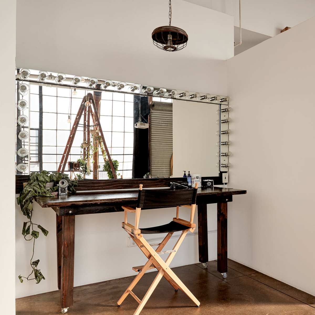 A minimalist makeup station featuring a large mirror with lights, a wooden chair, and a white partition in a well-lit room.