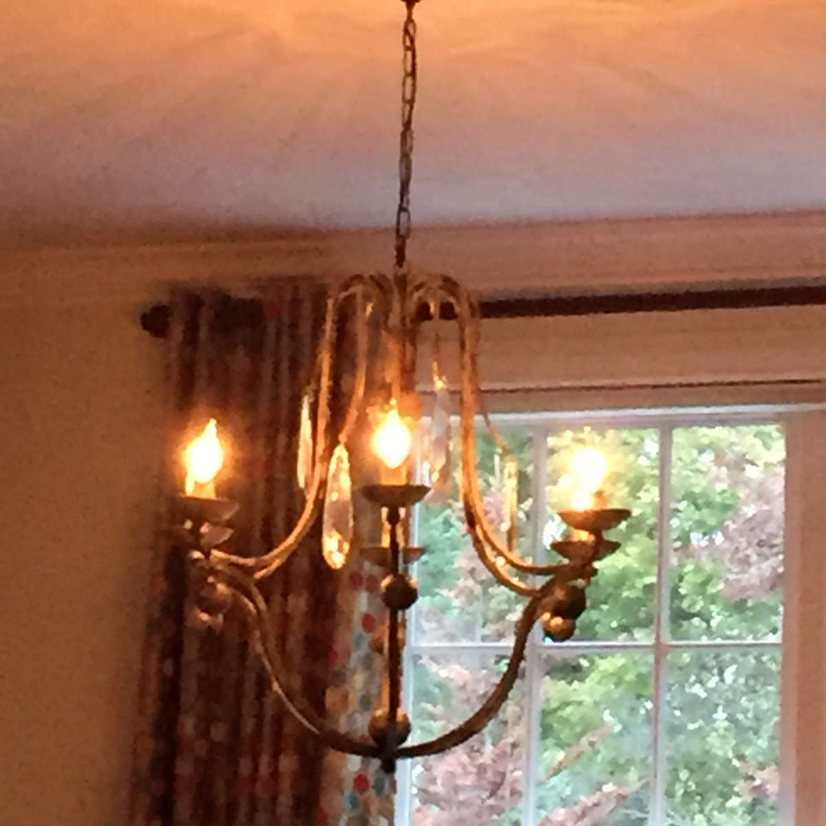 A chandelier with glowing bulbs hangs from the ceiling, casting light patterns on the wall in a room with a window and patterned curtains.
