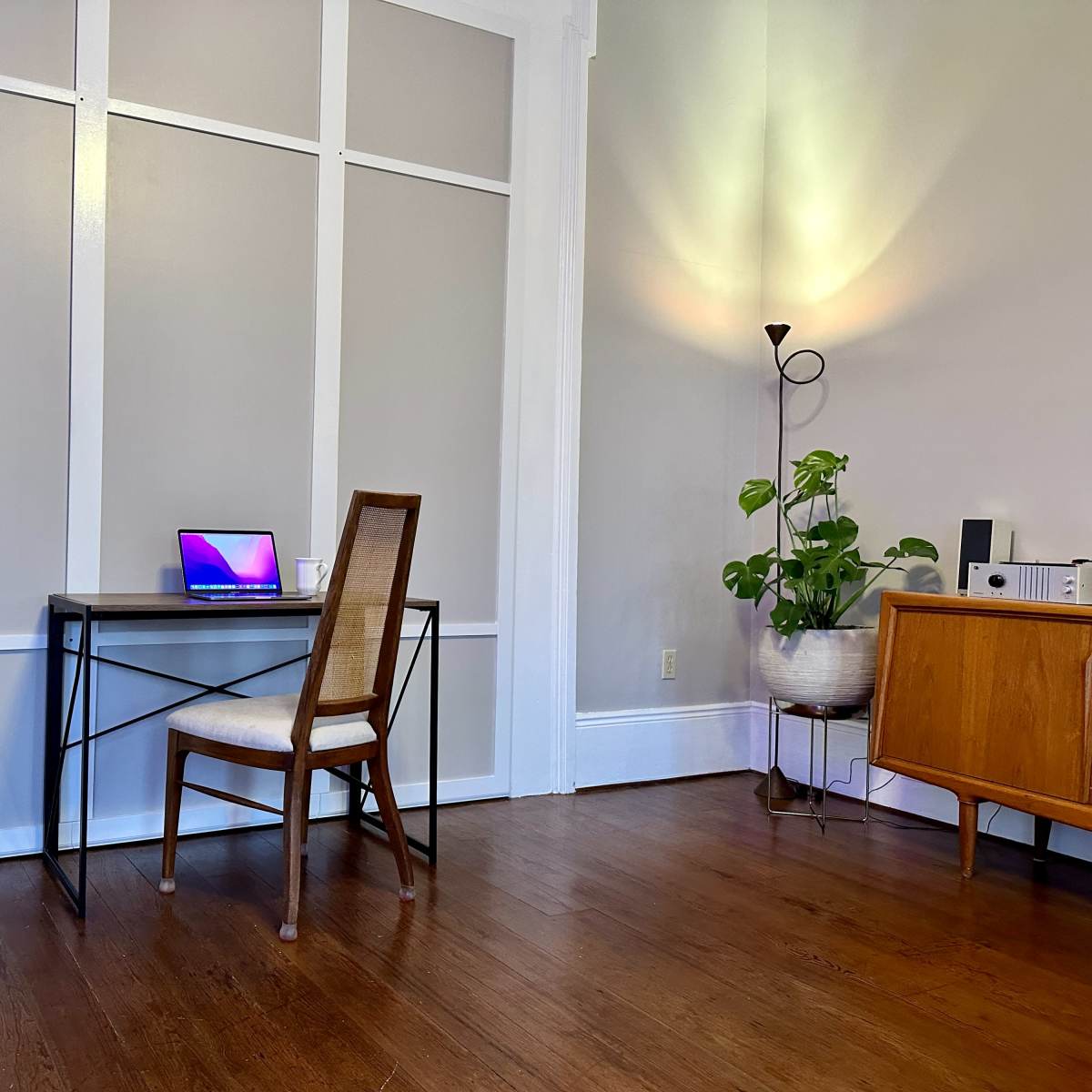 A simple desk with a laptop is positioned near a wall, accompanied by a chair and a potted plant in a well-lit room.