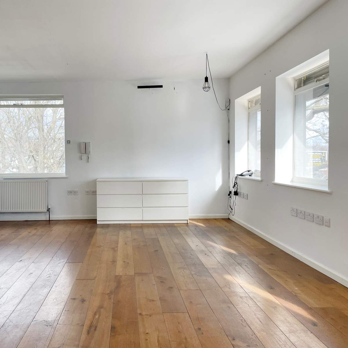 A spacious, empty room with light wood flooring and a white dresser against one wall, featuring large windows that allow natural light to fill the space.