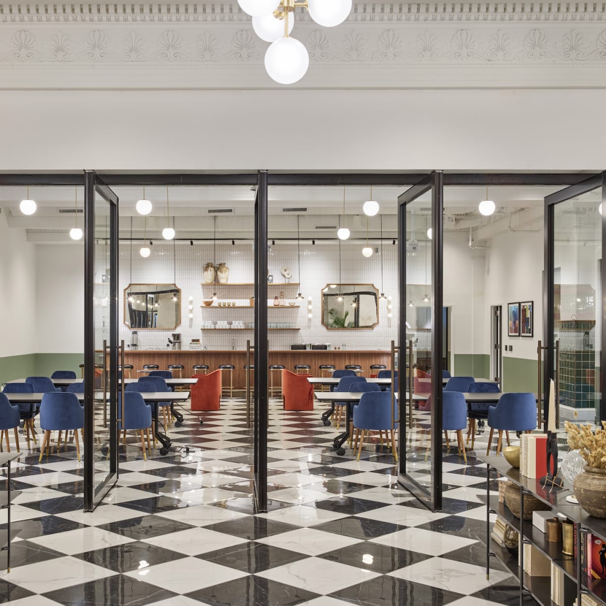 A modern interior space with a checkered black and white floor, featuring blue chairs and wooden tables, separated by glass doors from another area that includes decorative vases and artwork.