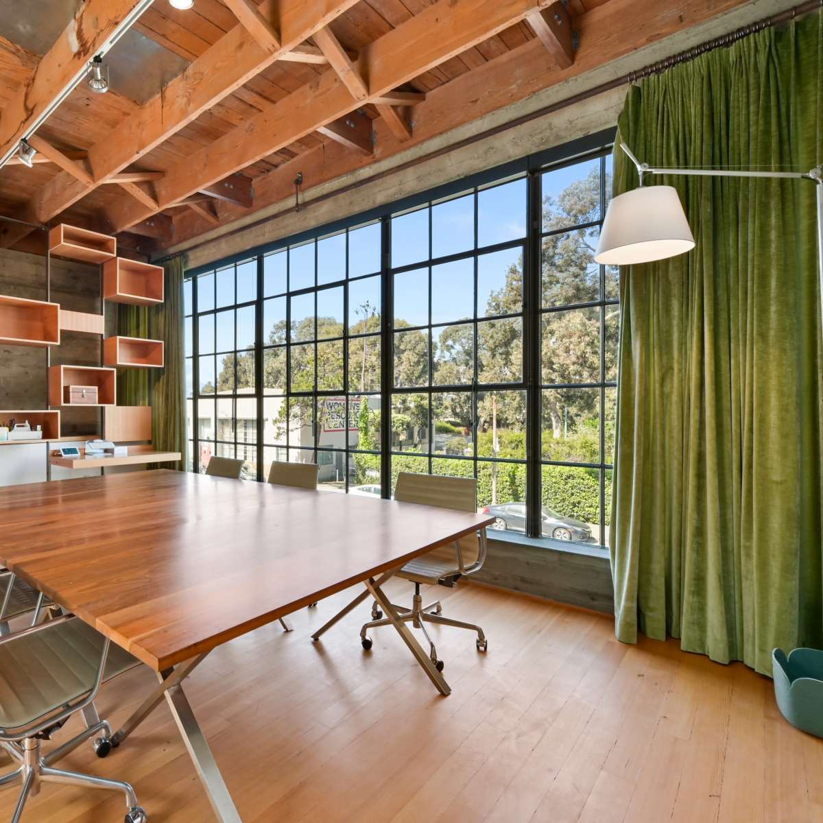 A modern conference room with a large wooden table, six metal chairs, and a large window flanked by green curtains, surrounded by shelving and a minimalist decor.