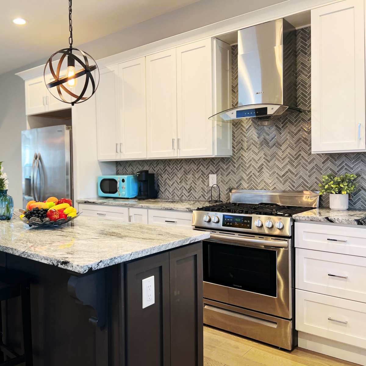 A modern kitchen with white cabinetry, stainless steel appliances, a gray and white herringbone backsplash, and a central island with bar stools and a fruit bowl.