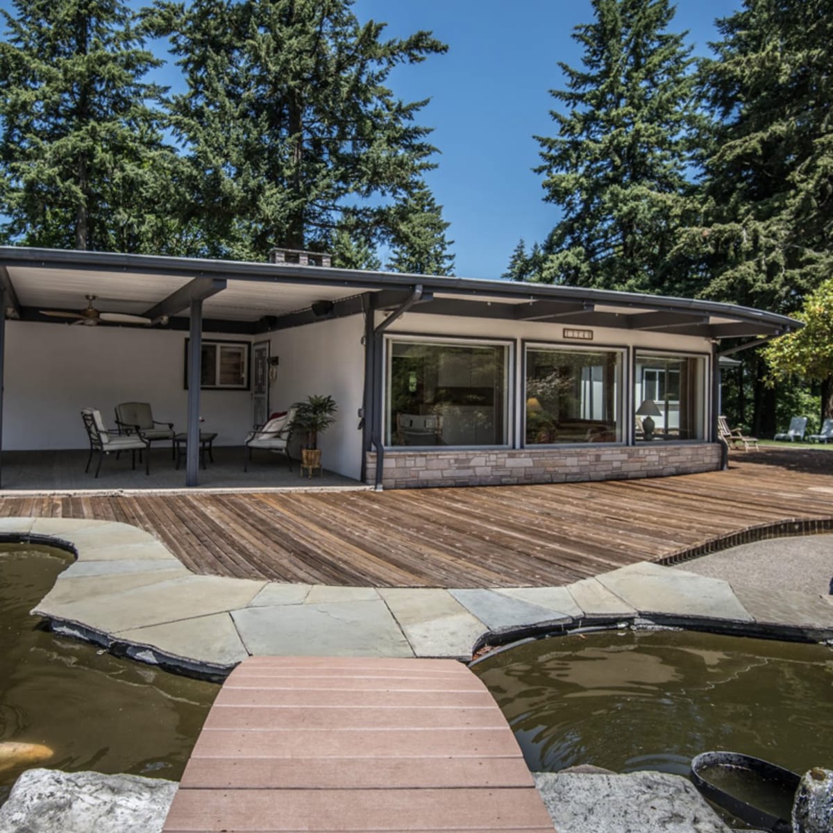 A modern house with a wooden deck, adjacent to a pond featuring koi fish, surrounded by tall trees.