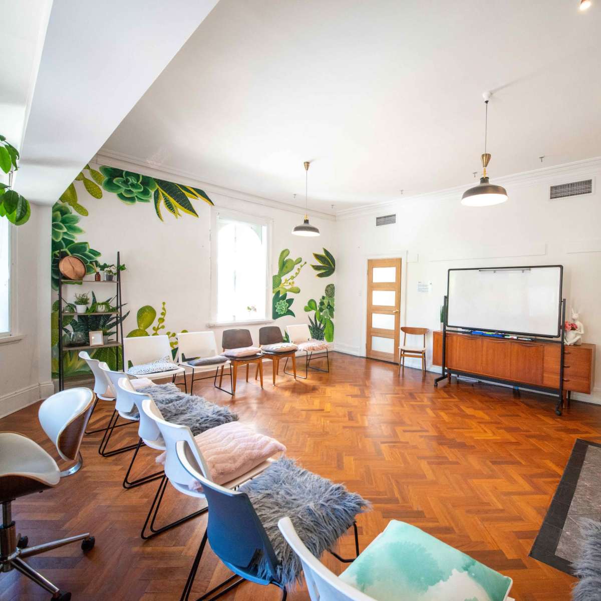 A spacious room with hardwood floors features a series of chairs arranged around a central area, a large whiteboard, and green plant murals on the walls.