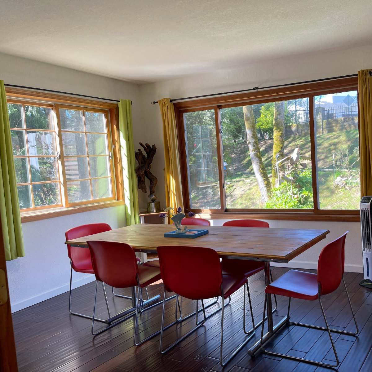 A dining area features a wooden table surrounded by red chairs, with large windows overlooking a green landscape.