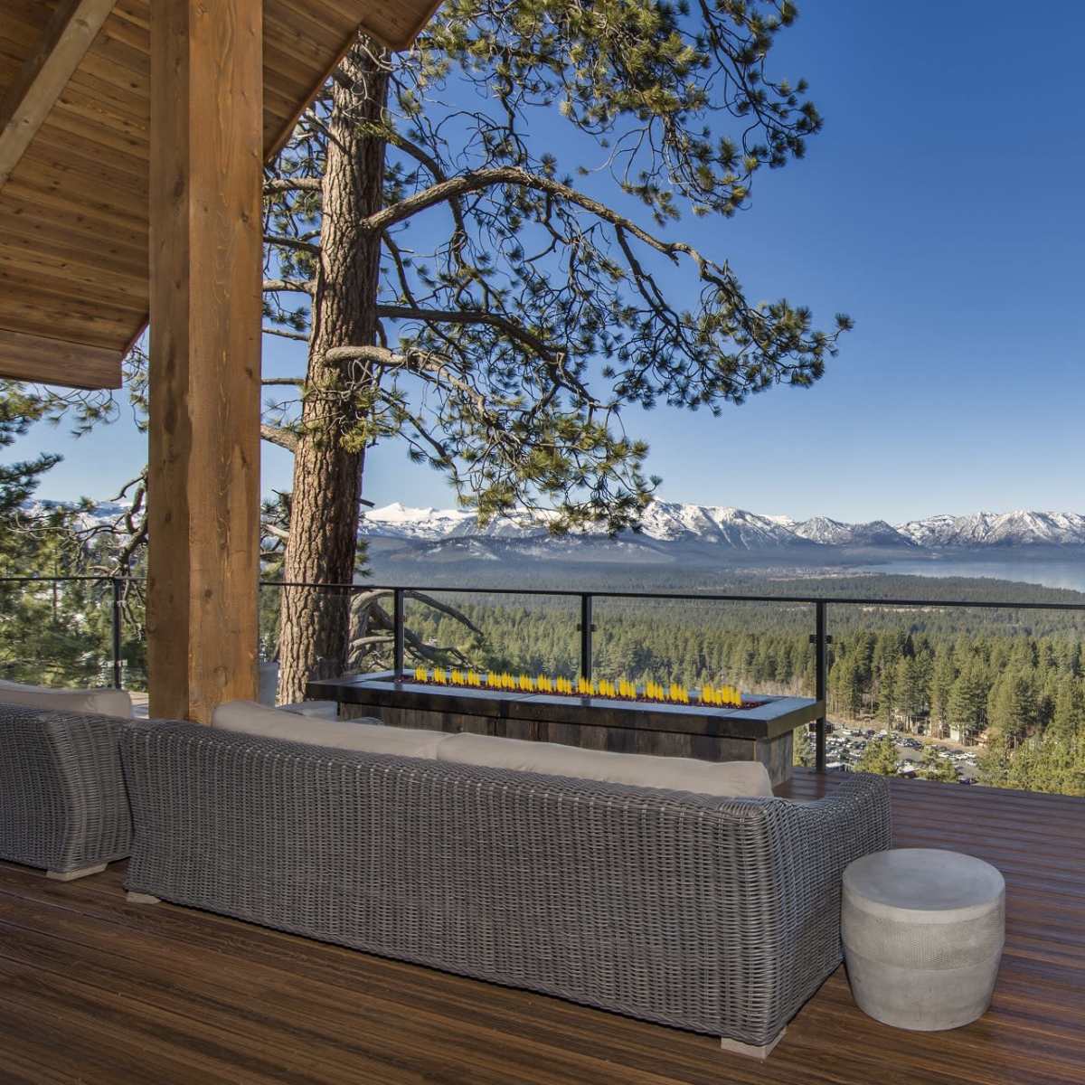 A spacious deck features comfortable seating and a fire table, overlooking a scenic view of mountains and a lake surrounded by trees.