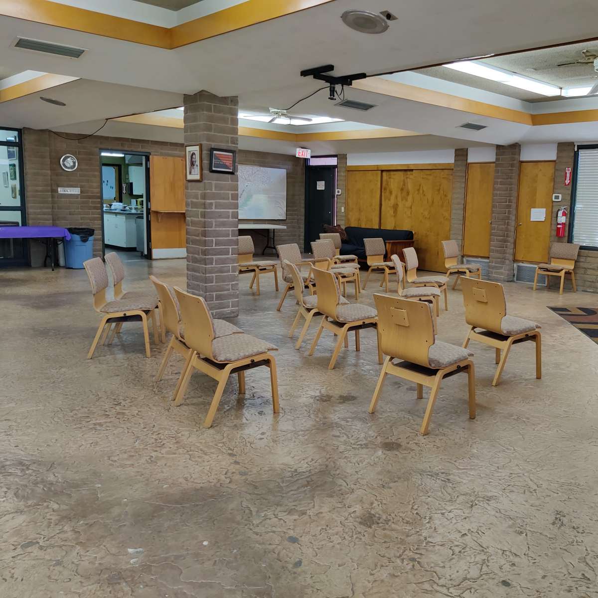 A room with several wooden chairs arranged in rows around a central space, featuring a podium with a cross and large windows along one side.