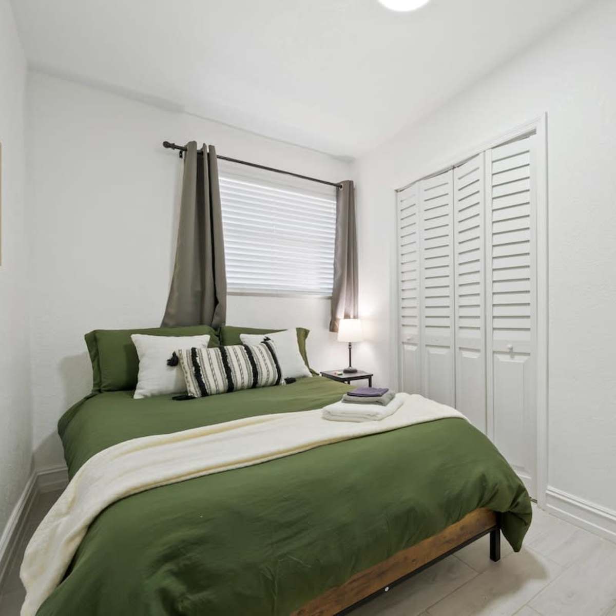 A small bedroom features a green bedspread, white pillows, and a nightstand with a lamp, complemented by a window with gray curtains.