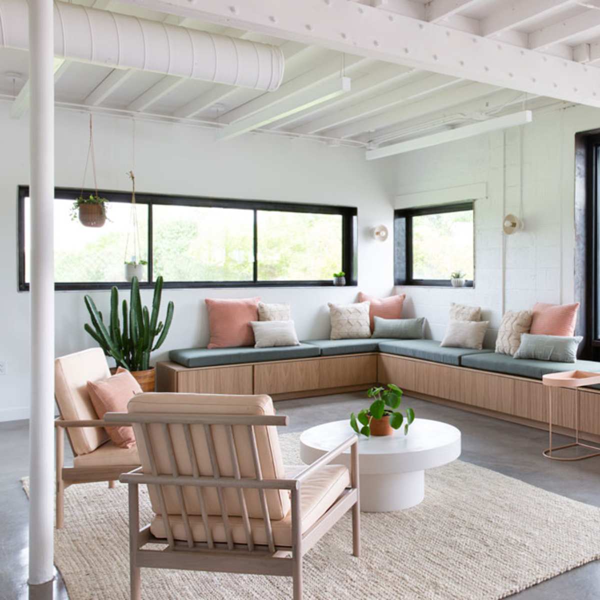 A bright and modern seating area features a large built-in bench with cushions, surrounded by windows and complemented by plants and a round coffee table.