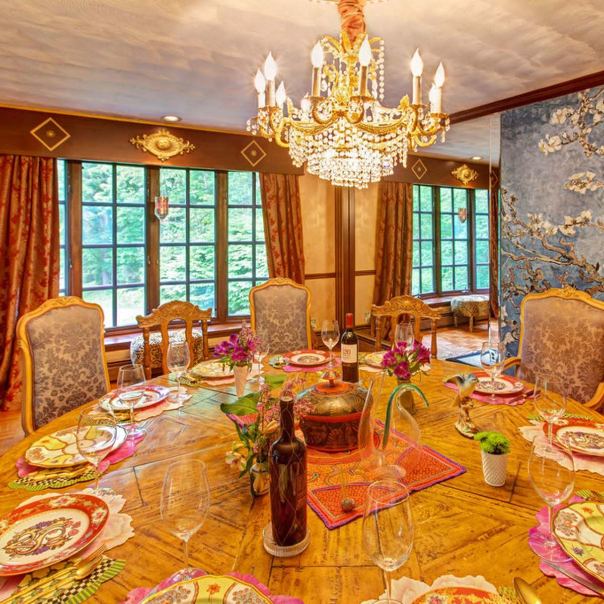 A dining room features a round wooden table set with colorful tableware and a chandelier hanging above, surrounded by ornate chairs and decorated walls.