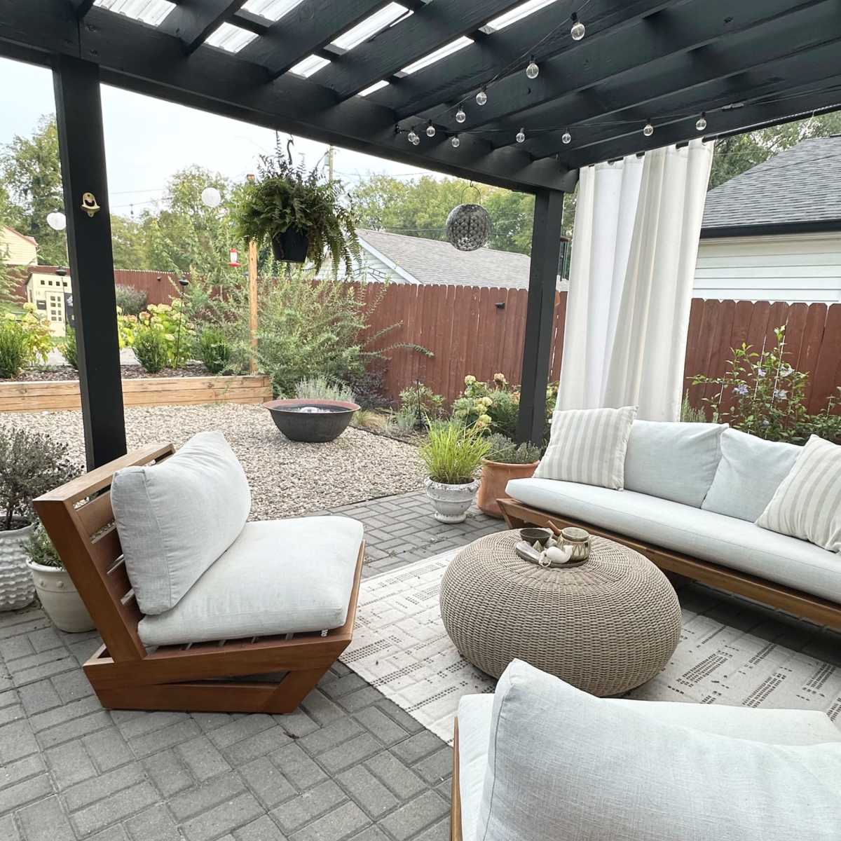 A cozy outdoor seating area with wooden furniture, a round pouf, and decorative plants under a shaded pergola.