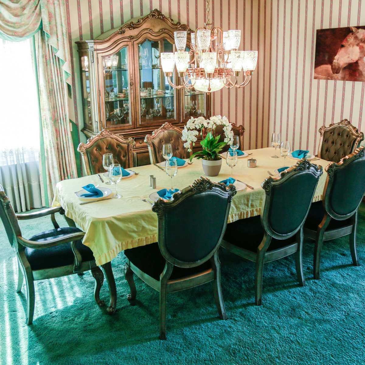 A formal dining room features a large table set for eight surrounded by ornate chairs, with a chandelier overhead and floral curtains framing a bright window.