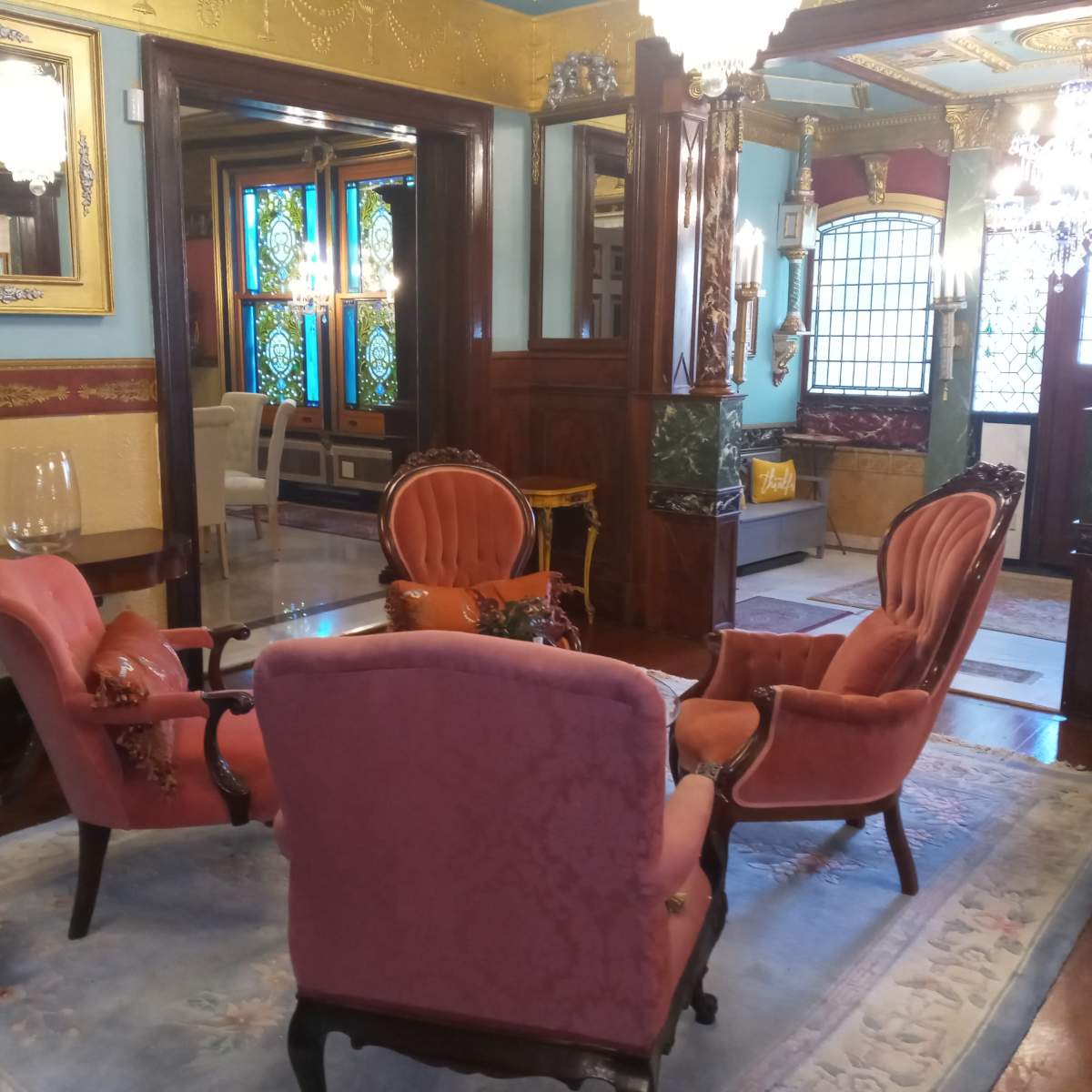 A vintage sitting area with four upholstered chairs arranged around a small table, surrounded by ornate decor and stained glass windows.