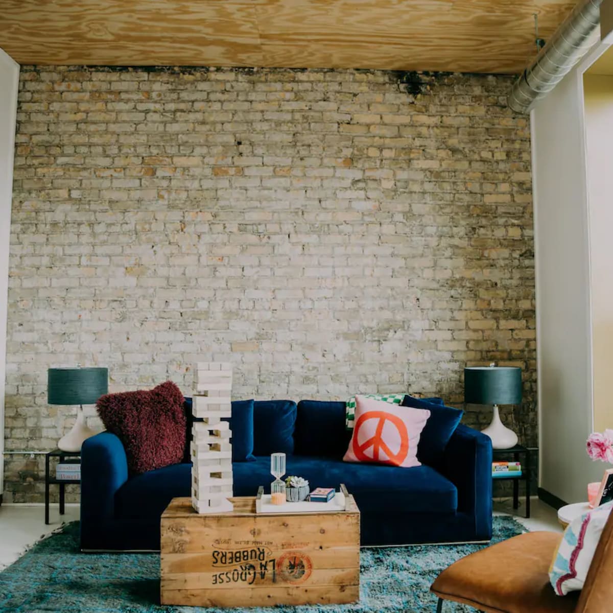 A cozy living room featuring a blue sofa, a wooden coffee table with games, and a decorative peace sign pillow, with exposed brick walls and large windows.