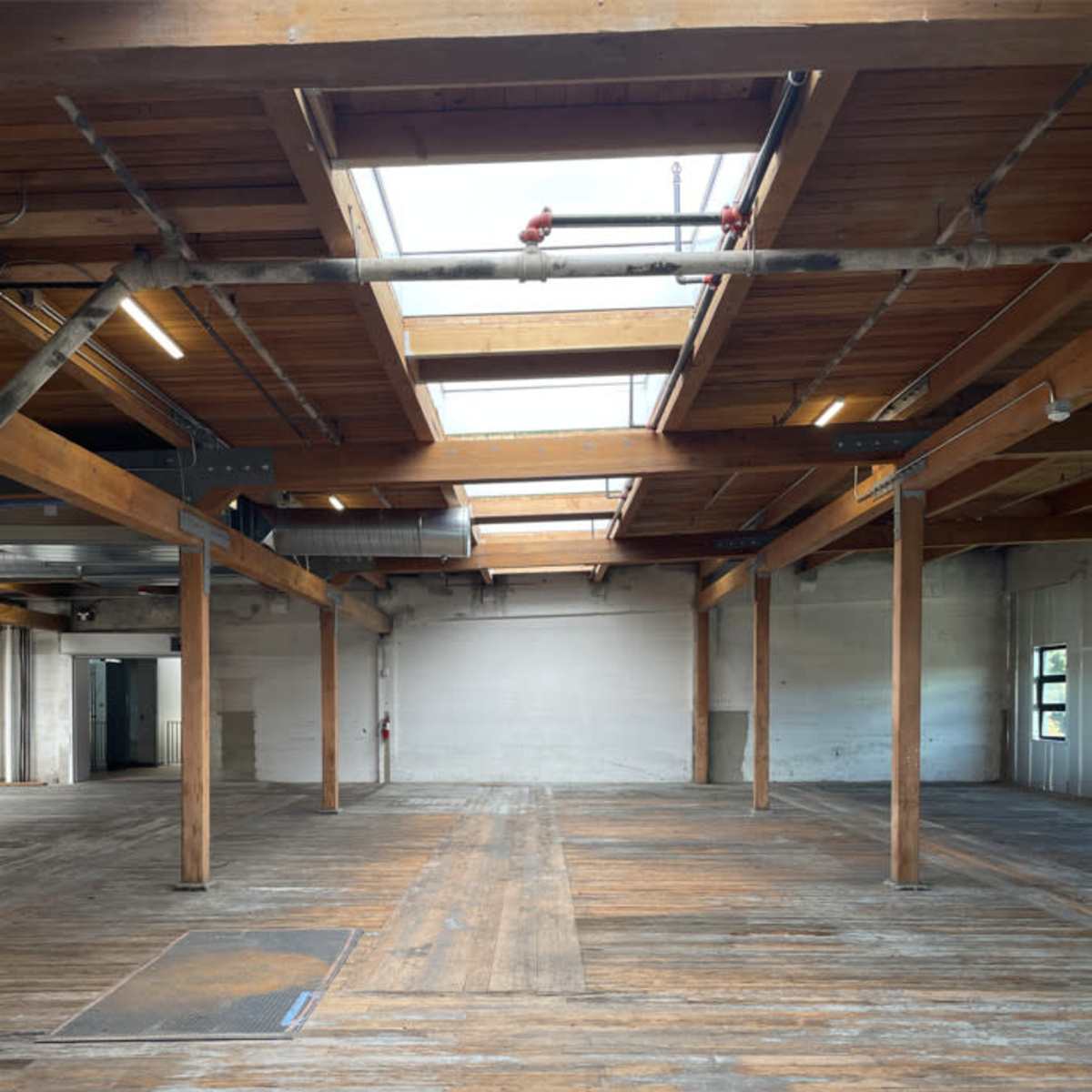 A spacious, empty room with wooden beams and a wooden floor, featuring skylights and several support columns.