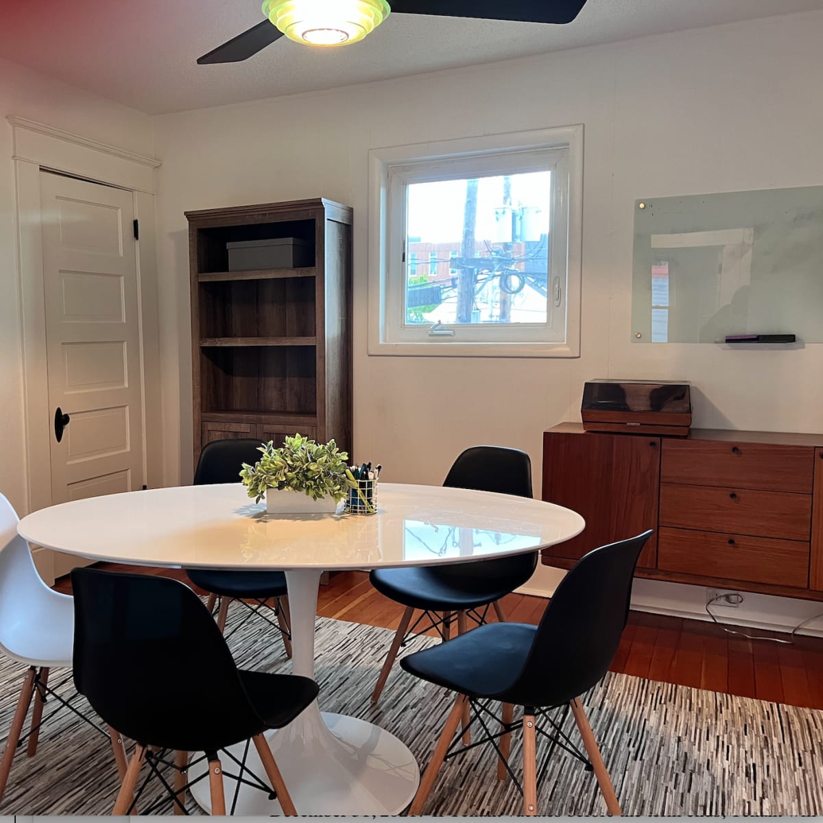 A round white dining table is surrounded by four chairs, with a plant centerpiece, in a well-lit room featuring a bookshelf and a wooden sideboard.