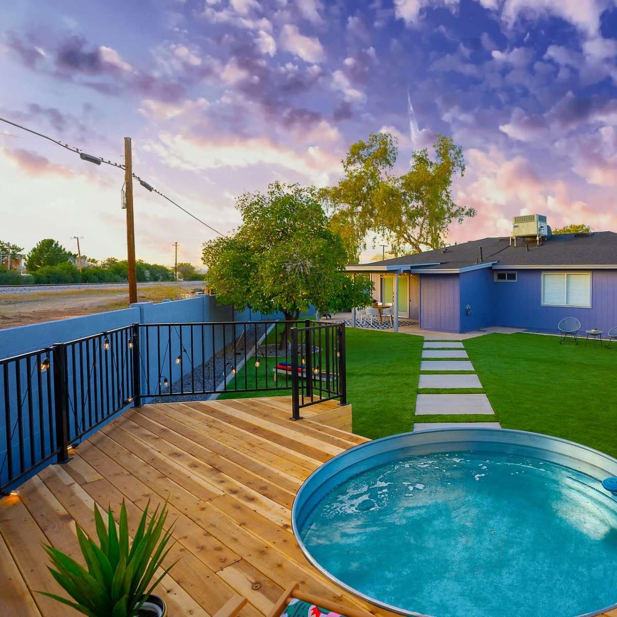 A wooden deck with a circular pool overlooks a grassy yard and a house painted purple, set against a colorful sky.