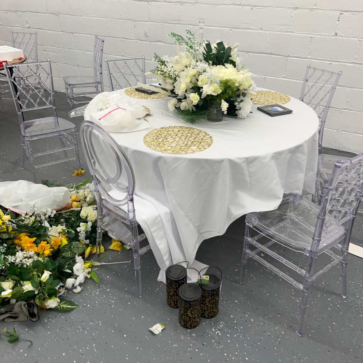 A round table covered with a white tablecloth is set with floral centerpieces and surrounded by clear chairs, while a pile of artificial flowers is scattered on the floor nearby.