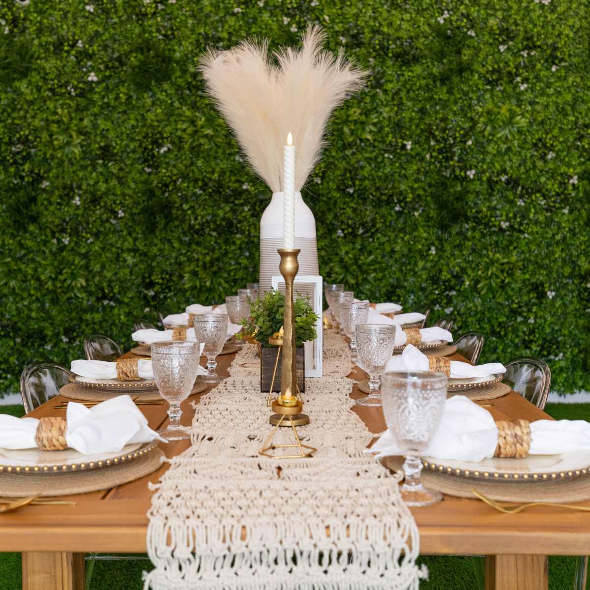 A long wooden dining table is set with white plates, clear glassware, and a central decorative arrangement on a textured runner, surrounded by a wall of greenery.