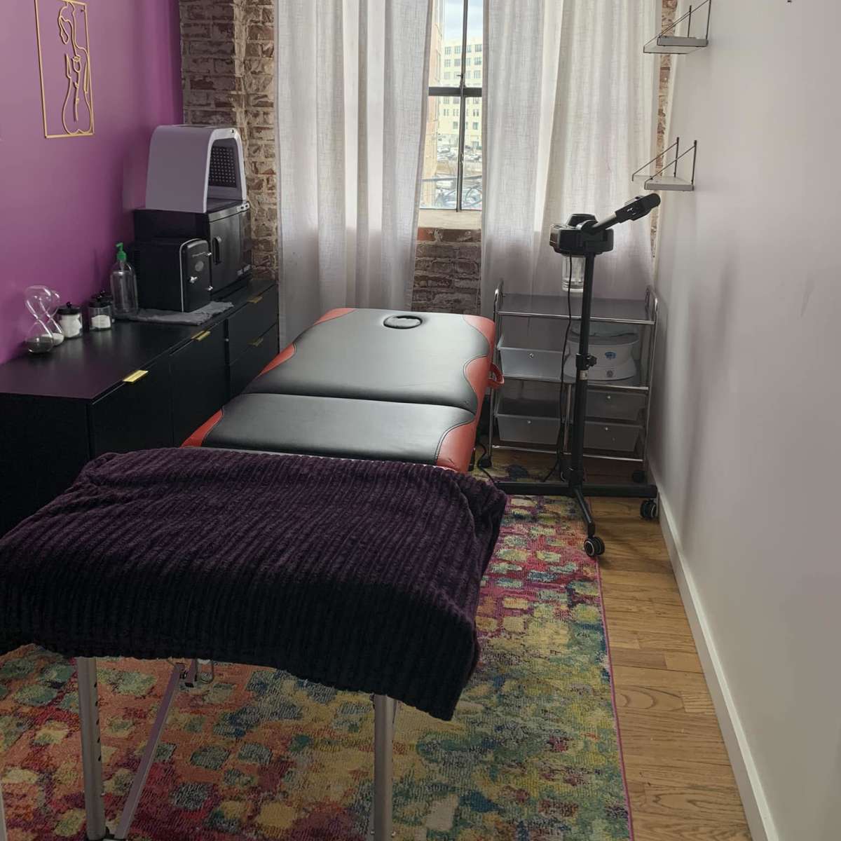 A treatment room with a massage table, a small storage cart, and large windows with sheer curtains.