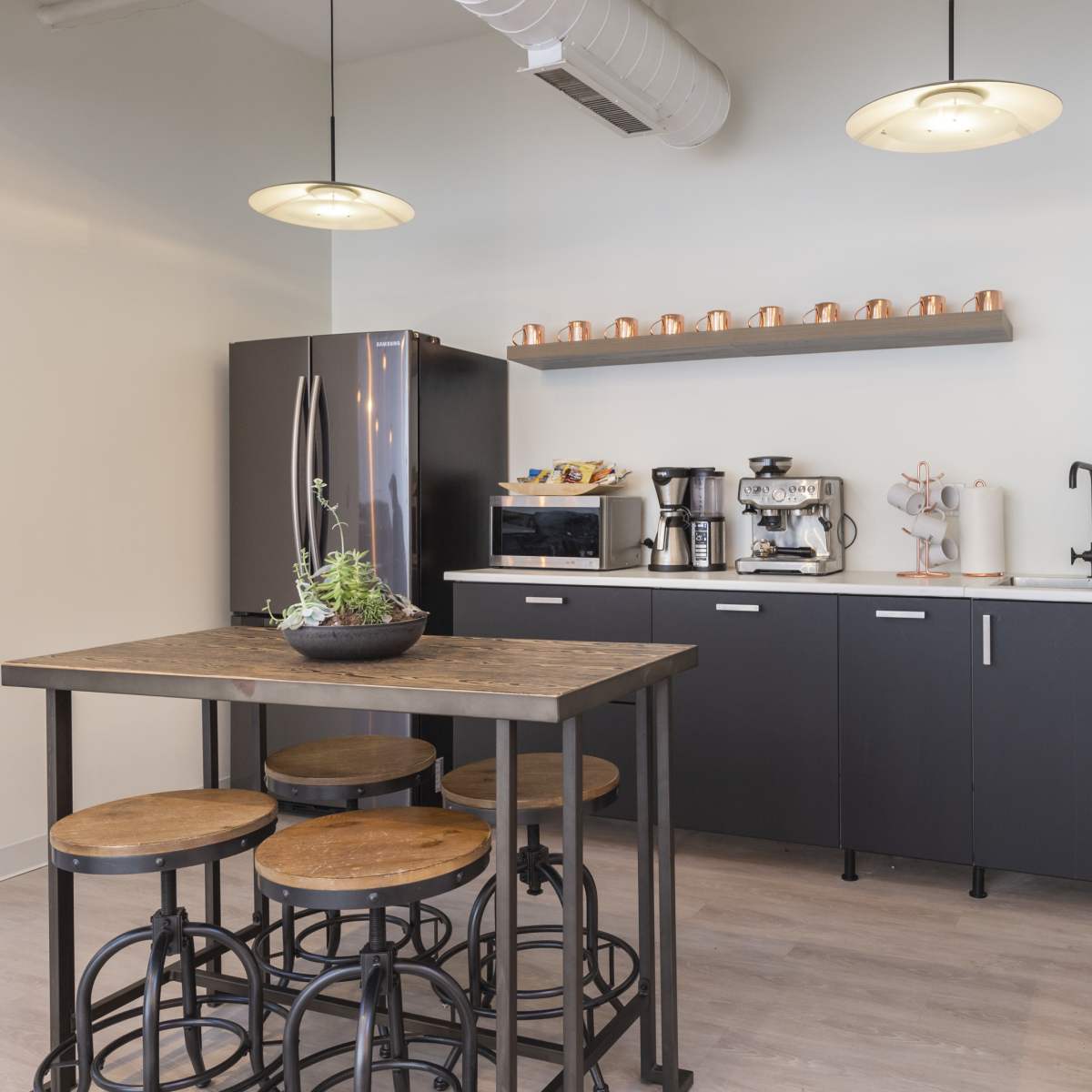 A modern kitchen area with a black refrigerator, coffee machines, and a wooden table surrounded by stools.