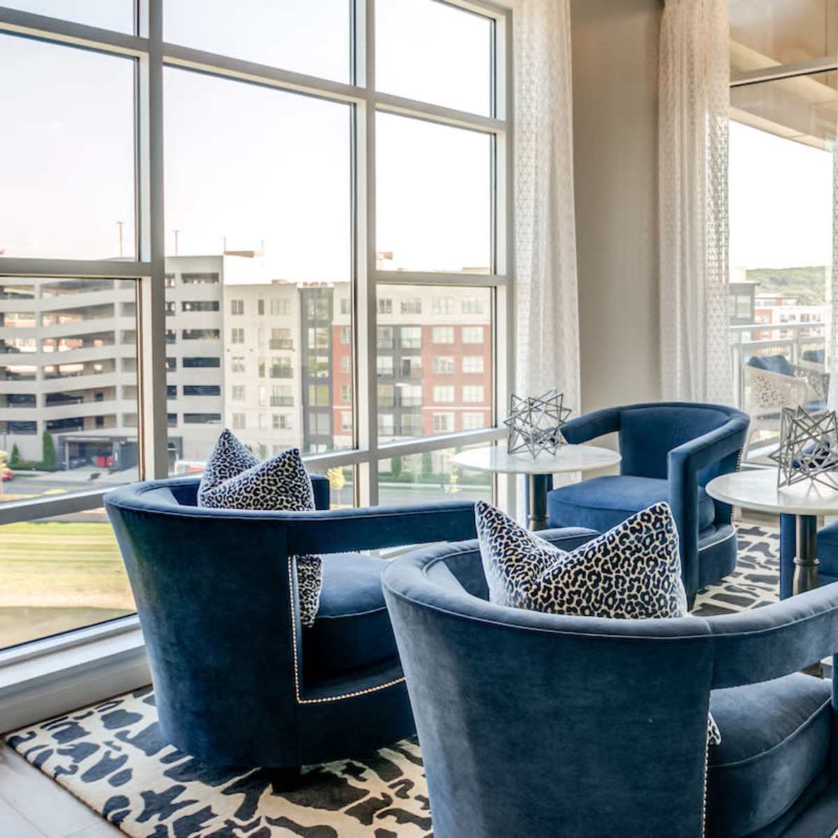 A stylish seating area features two blue armchairs with decorative pillows and glass-topped tables, surrounded by large windows overlooking a parking structure and nearby buildings.