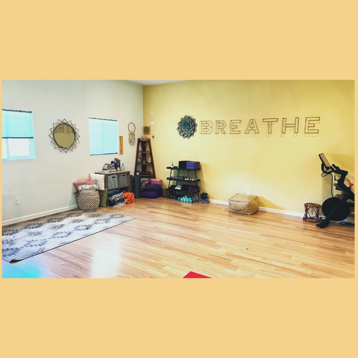 A spacious, well-lit yoga studio with a wooden floor, featuring a "BREATHE" wall decoration, yoga mats, and various props for exercise.