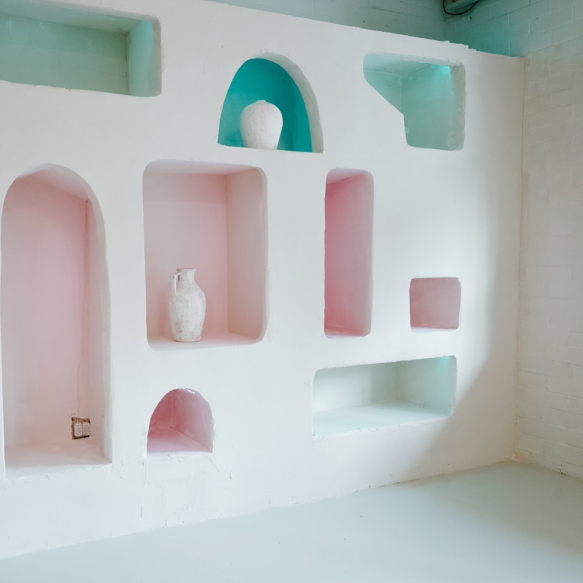 A white wall with various-shaped alcoves in pastel pink and teal, showcasing decorative vases.