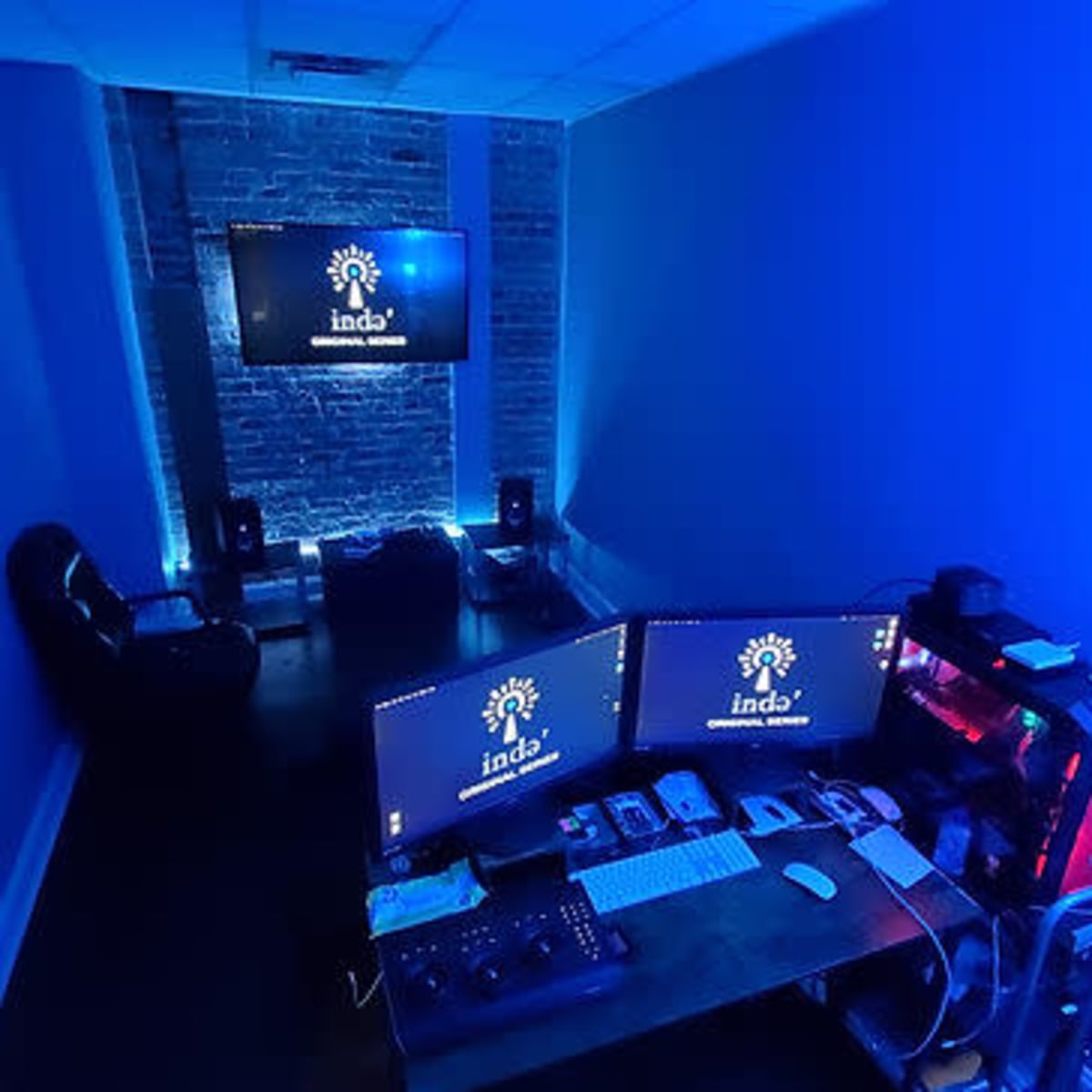A dimly lit office space with blue lighting, featuring two computer monitors, a desk with various devices, and a television on the wall.