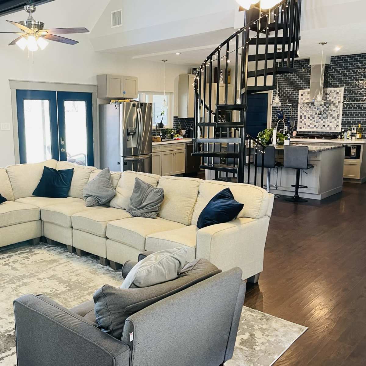 A spacious living area features a large sectional sofa with dark pillows, overlooking a modern kitchen with a spiral staircase in the background.