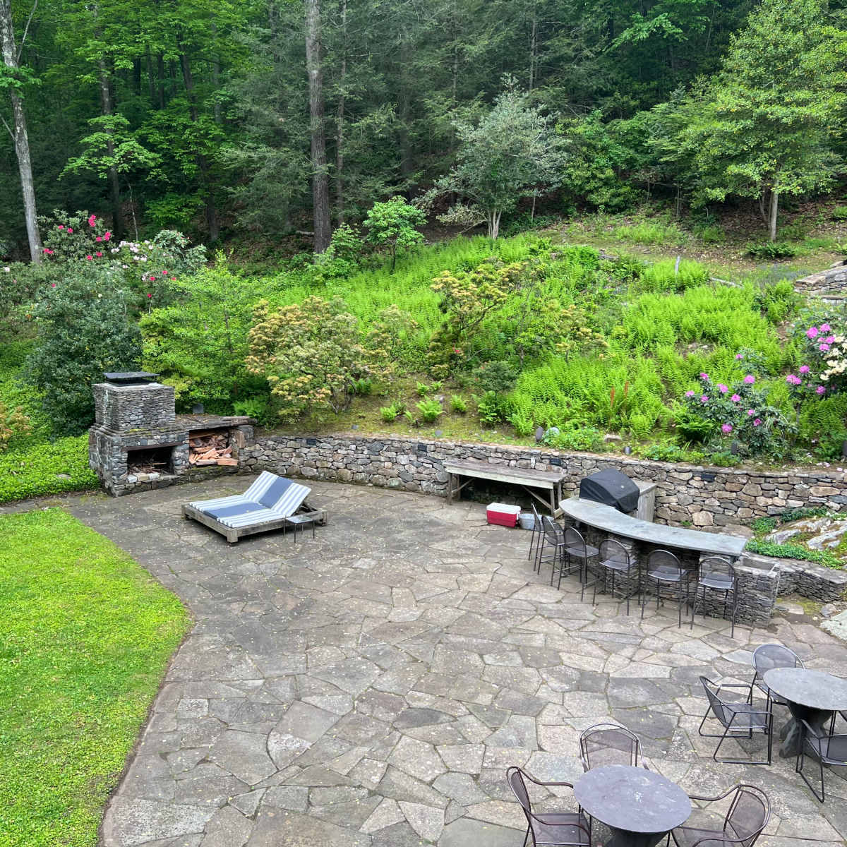 A stone patio area with lounge chairs, a fireplace, and a circular bar setup surrounded by lush greenery and flowering shrubs.