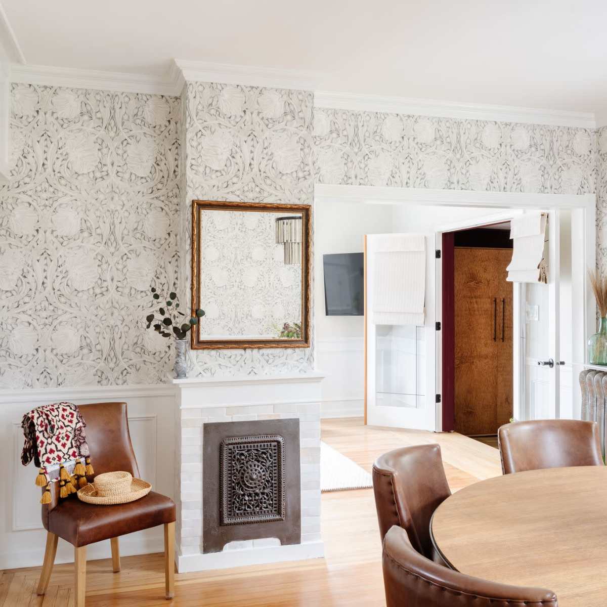 A dining area features a round wooden table surrounded by chairs, with a patterned wallpaper and a decorative mirror on the wall beside a vintage radiator.
