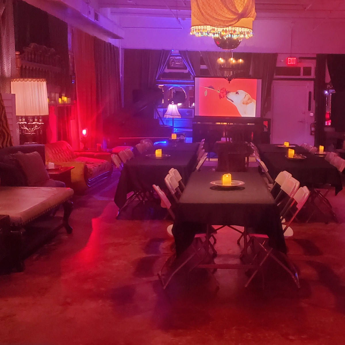 A dimly lit room with multiple tables covered in black cloth, surrounded by chairs, and illuminated by soft colored lights and candles, with a projector screen displaying an image at the far end.