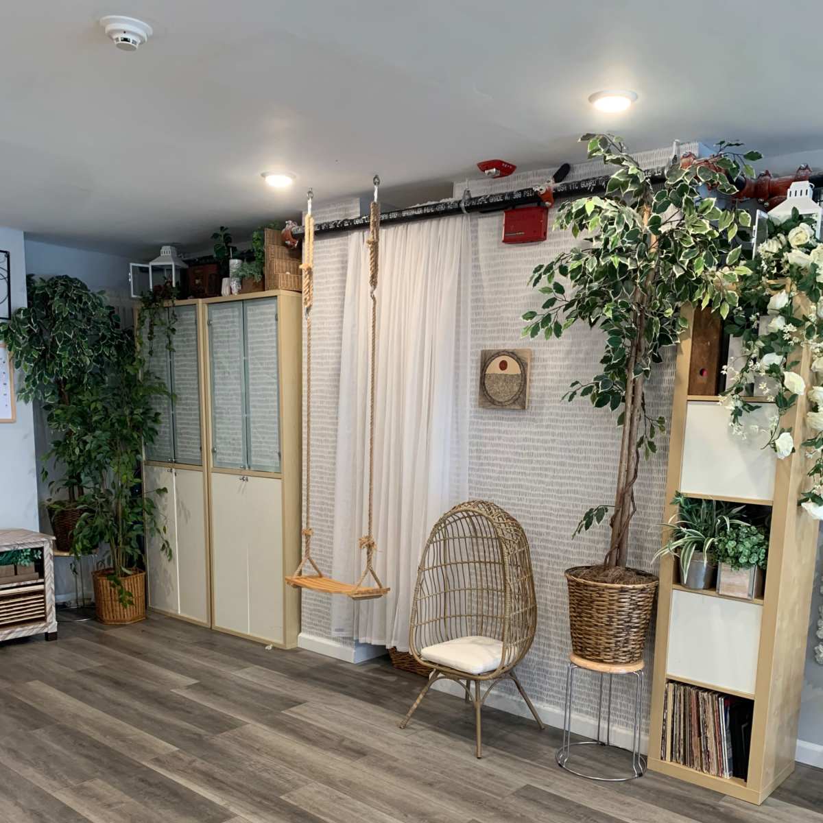 A cozy room with wooden flooring, a swing, plants, and shelves decorated with books and foliage.