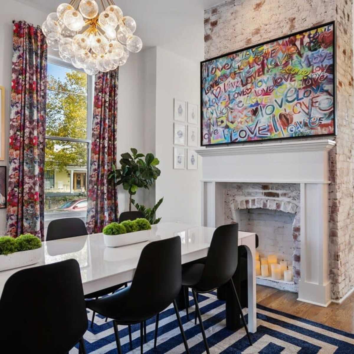 A modern dining area features a white table surrounded by black chairs, colorful curtains, and a large TV mounted above a brick fireplace.