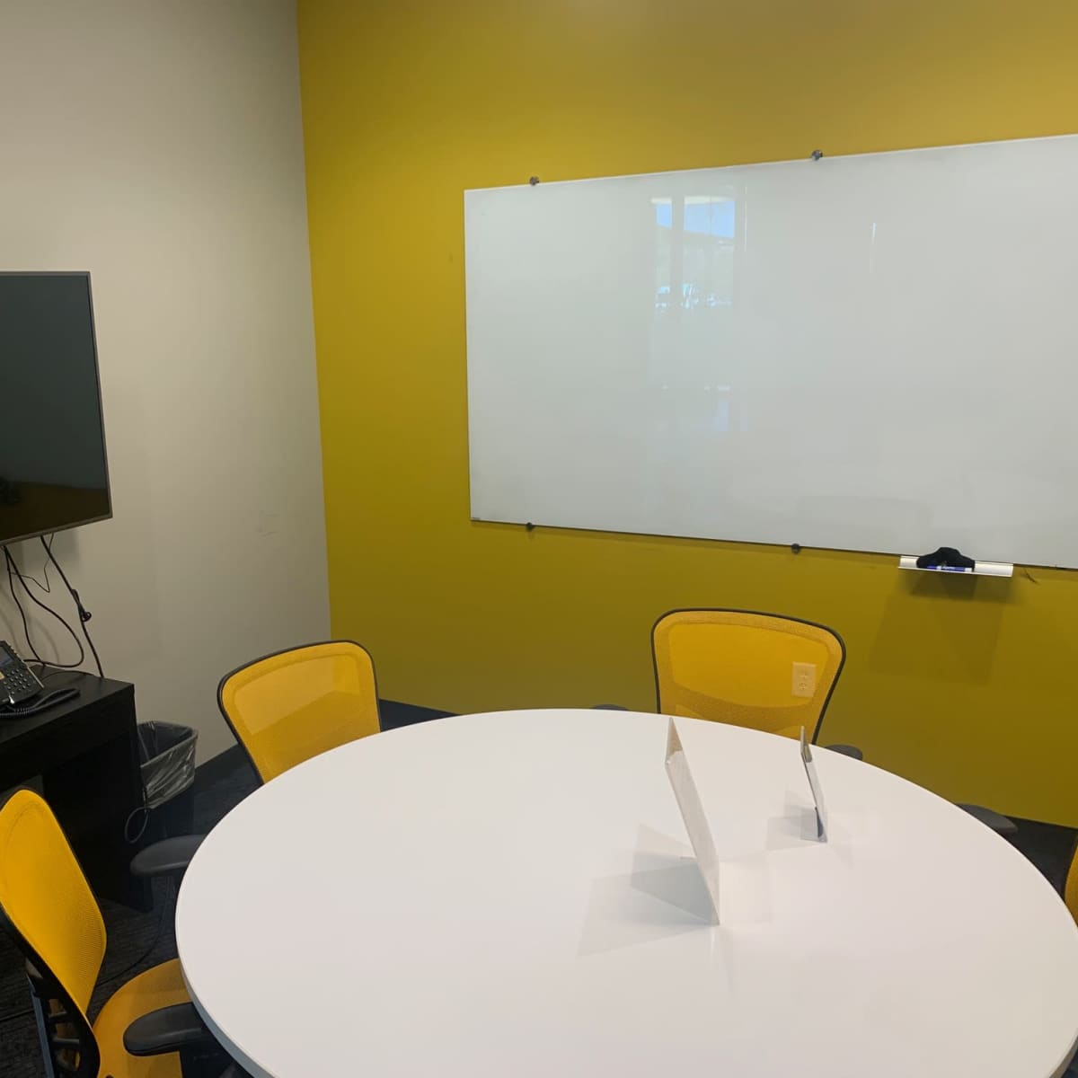 A conference room features a round white table, yellow chairs, a television mounted on the wall, and a whiteboard.