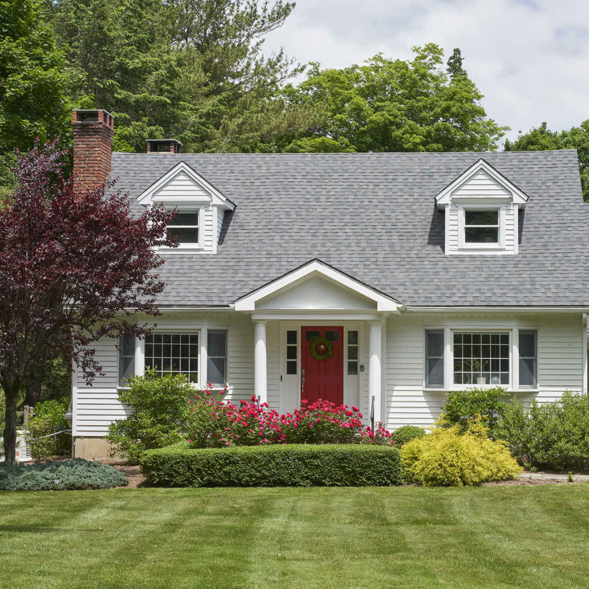 A two-story white house with a red front door is surrounded by landscaped greenery and flowering bushes.