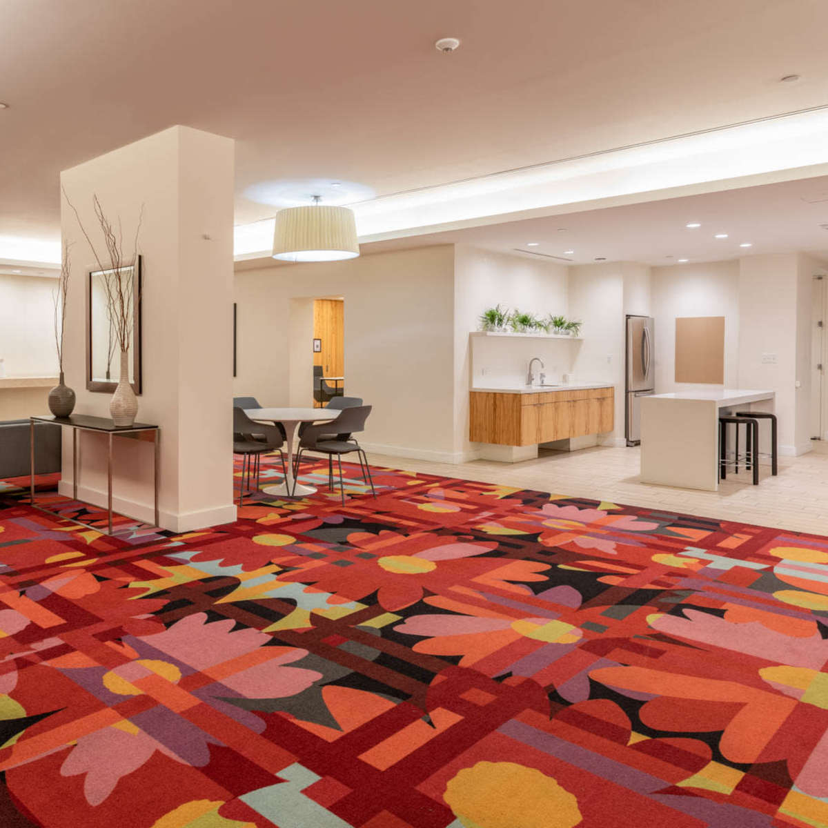 A modern lobby area with colorful floral carpet, seating arrangements, a television, and a kitchenette in an upscale interior space.